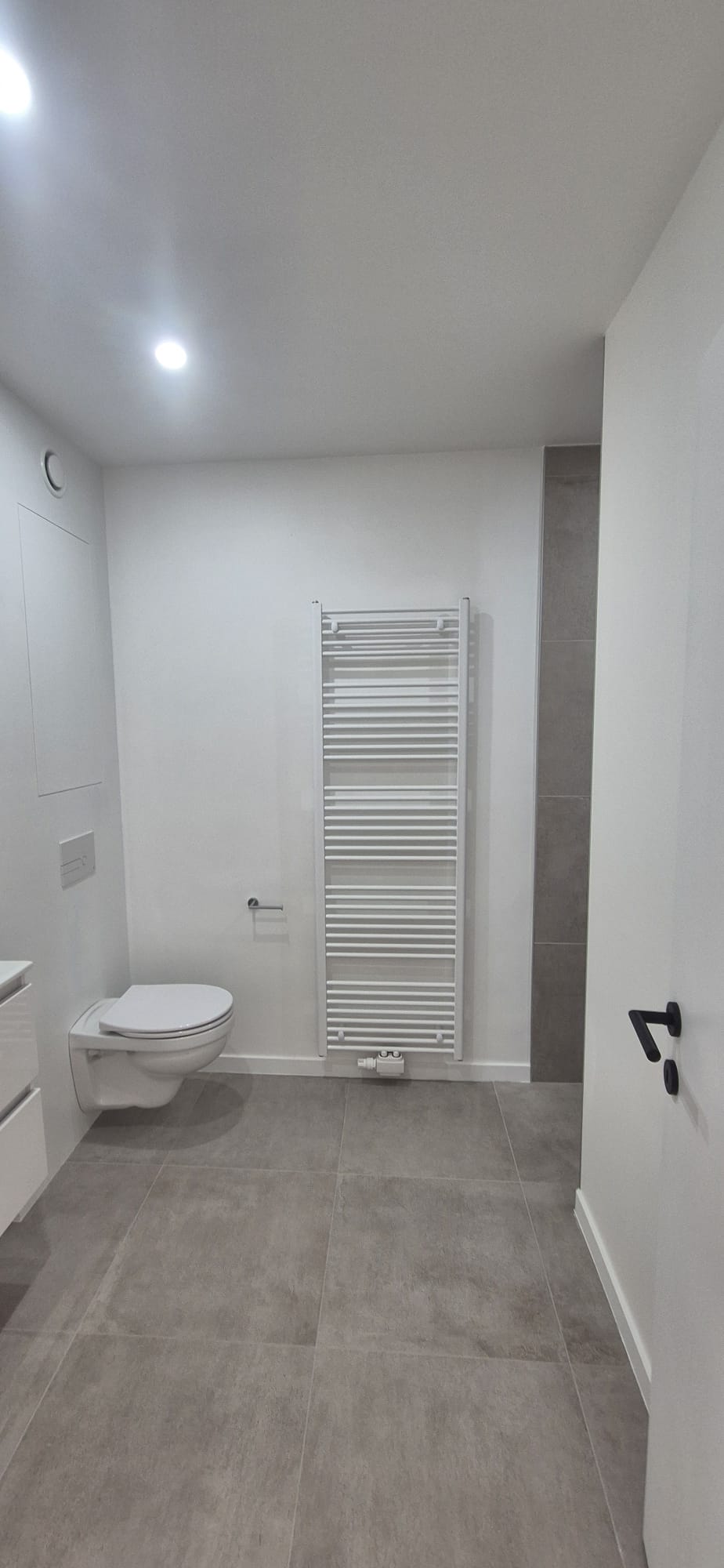 Moderne badkamer met hangtoilet, witte handdoekradiator en grijze tegelvloer — realisatie Artur Niko Ieper